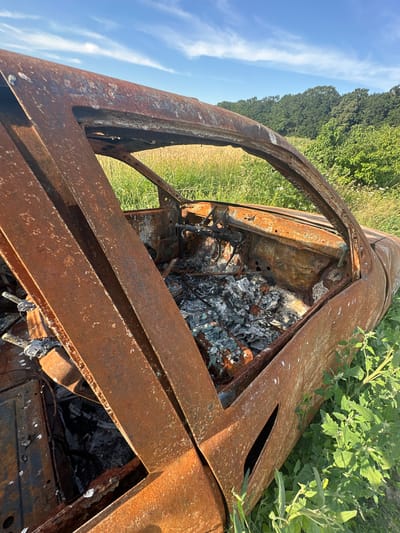 The media file shows the charred remains of a vehicle, likely a car, in an outdoor setting. The vehicle is heavily rusted and appears to have been burned, with ash and debris visible inside its interior. The scene is set in a rural or natural environment, with tall green grass and foliage surrounding the wreckage. In the background, a line of trees can be seen against a clear blue sky with some wispy white clouds. The perspective is from a high angle, looking down into the vehicle's interior and at its rusted metal components. The time of day appears to be daytime, with bright sunlight illuminating the scene. There are no people visible in the media file. The location context is Slobidka-Rakhnivska, Ukraine. No text is visible in the media file.