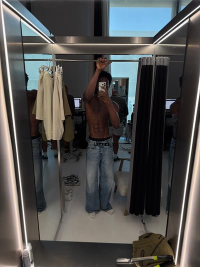 This media file depicts a full-length mirror selfie taken in what appears to be a fitting room or retail space. The primary subject is a fit individual, likely male, with visible abdominal muscles, posing shirtless and wearing light-wash denim jeans. They are holding a smartphone in their right hand, capturing the reflection.
In the background, the reflection reveals more of the setting. There are racks of clothing, including what appears to be beige and cream-colored tops or jackets hanging. A pair of athletic shoes is visible on the floor in the reflection. Other objects include black, stacked items, possibly folded textiles or materials, and what looks like a computer monitor on a desk in the distance. The overall aesthetic is modern and clean, with prominent lighting fixtures along the top and bottom edges of the mirror.
The lighting suggests it is daytime, as indicated by the natural light coming from a window or glass door visible in the upper left corner of the reflection. There are no overt emotional cues, but the pose and selfie format suggest a casual and perhaps self-aware moment.
The visible text on the jeans reads "DIESEL". Additionally, two cylindrical objects with labels are partially visible in the bottom right corner of the frame, near the edge of the mirror.