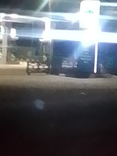 In a dimly lit outdoor environment, likely at night due to the artificial lighting present, a cart with four wheels is parked on an uneven, sandy surface. The cart is laden with multiple rectangular items stacked on top of each other, appearing to be sacks or boxes. The background features large, illuminated structures, possibly buildings or signage, with bright vertical and horizontal light sources. The overall scene suggests a commercial or storage area, with the cart and its contents indicating some form of transportation or staging of goods. The location is identified as BYE PASS, Nigeria.