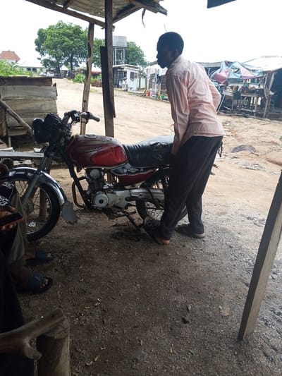 A man in a pale pink long-sleeved shirt and dark trousers stands next to a red and silver motorcycle under a wooden shelter. The motorcycle has the word "BOXER" visible on its fuel tank. The man is looking away from the camera, towards the right. The ground is covered in gravel and dirt. In the background, there are buildings, trees, and market stalls with awnings. The scene appears to be outdoors in a somewhat rustic setting, possibly a market or a workshop area. The overall lighting suggests it is daytime. The location is identified as Adankolo, Nigeria.
