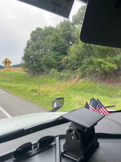 The image shows a view from inside a vehicle, likely a truck, looking out the windshield. In the foreground, on the dashboard, are a pair of black sunglasses and a black solar-powered device with an adjustable mount. Behind these objects, three small American flags are mounted on poles.
Outside the vehicle, the landscape consists of a grassy area with trees in the background. A yellow diamond-shaped road sign with black symbols indicating a railroad crossing and a road intersection is visible on the left. Below this sign is another rectangular yellow sign reading "TERRY FARM ROAD". A section of orange safety fencing is also present in the grassy area.
The sky is overcast, suggesting a cloudy day. The vehicle appears to be on a road, with a portion of the road visible to the left of the image. There are no people visible in the image. The overall scene depicts a roadside view from a vehicle, possibly in a rural or semi-rural area. The location is identified as Arcade, United States of America, based on the sign reading "TERRY FARM ROAD".