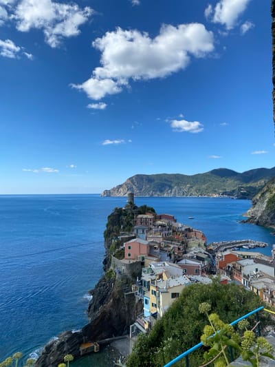 The image displays a high-angle, wide-perspective view of Vernazza, Italy, a coastal village situated on a rugged promontory extending into the Mediterranean Sea. The scene is illuminated by bright sunlight under a clear blue sky with scattered white cumulus clouds.
The village of Vernazza occupies the central-right portion of the frame, characterized by densely packed, multi-story buildings with colorful facades in shades of pink, yellow, orange, and terracotta. The cylindrical stone tower of Doria Castle stands atop the highest point of the promontory, visible to the left of the main cluster of buildings. To the right of the village, a small harbor contains several white boats docked within a stone-walled basin.
The deep blue Mediterranean Sea dominates the left and middle ground, with a single small white vessel visible in the open water to the left of the promontory. A faint line, possibly a boat wake, extends across the water further left. Distant green, forested mountains and hills line the coastline in the background, curving towards the right. The immediate foreground features green foliage and a partial blue metal railing. No identifiable individuals or specific actions are discernible within the frame.