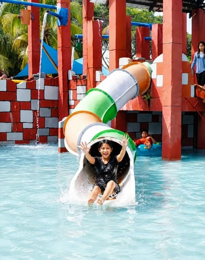 A young girl with dark hair and a black shirt is pictured on a water slide at a water park in Pantai Cermin Kiri, Indonesia. She is mid-slide, with water splashing around her as she emerges from the white tube. Her arms are raised in the air and she has a wide smile on her face, appearing joyful. The water slide itself is multicolored, with sections of orange, green, and white. In the background, red pillars and red and white checkered walls suggest the structure of the water park. Further back, another child is visible sitting in a blue inner tube in the water. The water in the pool is a clear, bright blue. The environment is sunny, with palm trees visible in the upper background, indicating a tropical location.