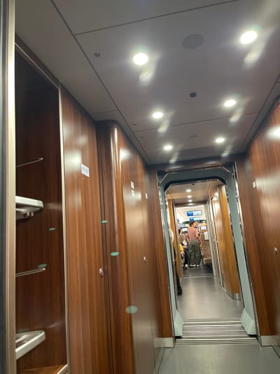 This media file captures a perspective looking down into a modern, multi-level interior, possibly a train or a sophisticated elevator shaft. The dominant materials are polished wood paneling and sleek metal accents, suggesting a high-end design.
In the distance, at the lower level, two individuals are visible. One person, wearing a striped shirt and green pants, is standing and looking towards the camera. Beside them, another person appears to be seated. Various bags are placed around them, indicating travel or a waiting period.
The lighting in the scene is a mix of bright, circular overhead lights and the warm glow reflecting off the wood surfaces. The overall ambiance is clean and contemporary. The view down the shaft reveals multiple levels of wooden surfaces with subtle metallic trim. Some of the wooden surfaces have small white stickers on them.
The image appears to be taken indoors, likely during the day given the bright illumination. The specific location context provided is Longhua District, China, which could imply this is a transportation hub or a well-designed public space within that region. The reflection of lights on the polished wood creates a sense of depth and shimmer throughout the space. The metallic elements, particularly the railings and vertical grooves on the far wall, add a touch of industrial elegance. The scene conveys a sense of order and advanced design.