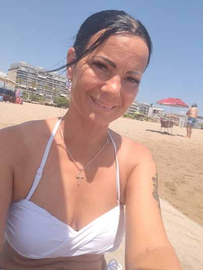 A woman with dark hair pulled back and wearing a white bikini top and a cross necklace poses for a selfie on a sandy beach in Calafell, Spain. She is smiling broadly, and her makeup is visible. Her left arm, bearing a tattoo, is resting on a beige mat. In the background, there are buildings, palm trees, beach chairs, umbrellas, and other beachgoers, including a man in blue shorts standing near a red umbrella. The sky is clear and blue.