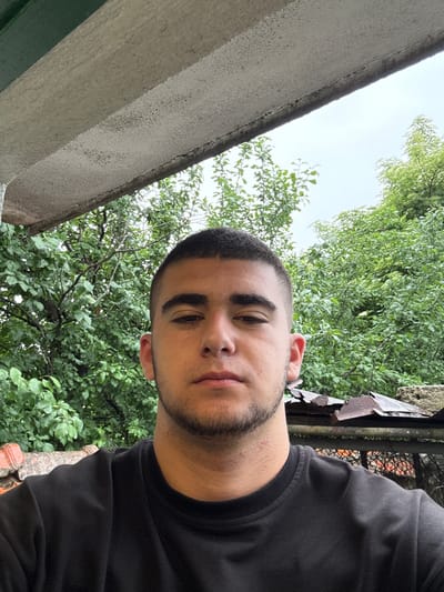 The image captures a close-up, slightly upward-angled portrait of a young man outdoors in Krushare, Bulgaria. The primary subject is a male with short, dark hair and light facial stubble, wearing a plain black t-shirt. He is looking directly at the camera with a neutral to slightly pensive expression.
The setting is characterized by lush green foliage, likely trees or bushes, which fill most of the background. To the left, a light-colored, rough-textured concrete or stucco structure, possibly part of a building or a retaining wall, extends diagonally upwards. Its surface shows some wear and irregular texture. On the right side, behind the man's shoulder, there are hints of a weathered and somewhat damaged structure, possibly metal sheeting or a fence, with glimpses of a reddish-brown object or wall through its openings. The sky, visible between the foliage and the structure, appears bright and overcast, suggesting a cloudy daytime. No specific activity beyond posing for the photo is evident. There is no visible text in the image.