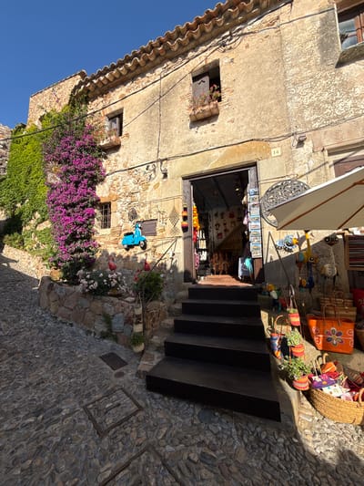 The image depicts a charming, narrow cobblestone street in Tossa de Mar, Spain, bathed in bright sunlight, suggesting it's midday. The scene is dominated by an old stone building with a weathered facade, adorned with vibrant purple bougainvillea and lush green ivy cascading down its walls.
In the center, a small shop with an open doorway showcases a variety of colorful crafts and souvenirs, including woven bags, pottery, and what appear to be decorative figurines. A white umbrella is propped open above the shop entrance, providing shade. Dark wooden steps lead up to the shop from the street.
Notable details include a small, bright blue decorative scooter mounted on the wall near the shop, and several colorful woven baskets and bags displayed outside. A sign with text, partially obscured by flowers, is visible on the wall. The overall atmosphere is idyllic and tranquil, evoking a sense of old-world charm and Mediterranean beauty. The clear blue sky indicates fair weather.