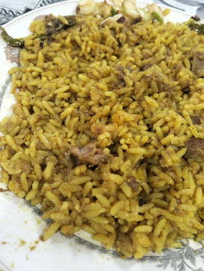 A close-up shot of a plate of meat biryani. The rice is yellow, possibly due to turmeric or saffron, and mixed with visible pieces of meat. There are green chilies and what appear to be pieces of cucumber and other vegetables on the edge of the plate. The plate itself is white with a gray floral border. The location is Naodoba, Bangladesh, as indicated in the prompt.