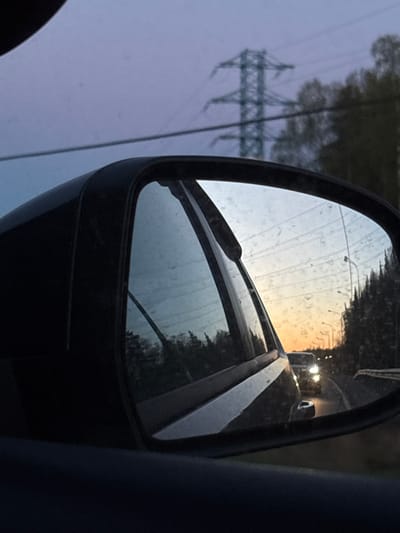 The image depicts a car's side mirror reflecting a road and surrounding environment. The mirror shows a roadway with at least one other vehicle with headlights visible in the distance. Trees are visible along the side of the road in the mirror. A utility pole is visible on the left side of the image, out of the mirror's reflection. The image appears to have been taken at dusk or dawn given the lighting. The location is inferred from the prompt as Myaglovo, Russia. There are no people visible in the image.