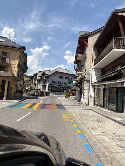The image depicts a street scene in Saint-Gervais-les-Bains, France, on a sunny day with scattered clouds. The street features a vibrantly colored pedestrian crossing with stripes of teal, yellow, and purple. The road is paved and marked with white lane lines. To the right of the road, a sidewalk is constructed with light-colored paving stones, bordered by blue and yellow painted markings.
Several buildings line the street. On the left, a building with a light yellow facade has balconies and shutters. Further down the street, more buildings with traditional alpine architecture, featuring wooden balconies and pitched roofs, are visible. One building on the right side of the street has a sign that reads "Pâtissier Fournil du Centre," indicating a bakery. Another building features a prominent green cross, suggesting a pharmacy.
A person wearing a backpack is standing on the sidewalk on the left side of the street. The hood of a dark-colored car is visible in the foreground, suggesting the image was taken from inside a vehicle. The overall environment appears to be a town center, likely a tourist area given the architecture and the presence of shops and amenities. In the background, a hillside with greenery is visible, suggesting the mountainous terrain surrounding the town.