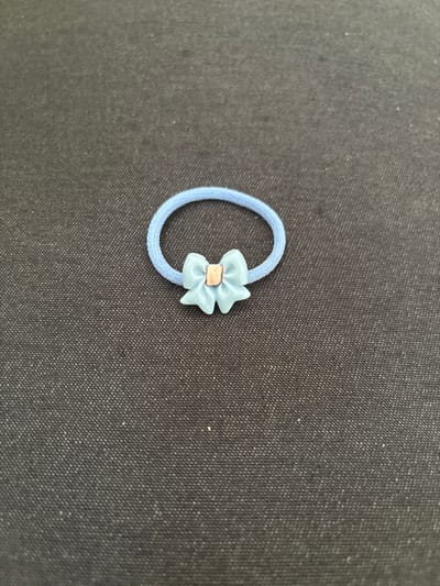 A light blue, elastic hair tie with a small, light blue bow attached to it rests on a dark, textured fabric surface. The bow has a light pink or tan square accent in its center. The background fabric appears to be a dark denim or canvas material with a visible weave pattern. The scene is evenly lit, and the object is centrally located. The image does not depict any elements that would allow for the determination of its location in Imeni Morozova, Russia, as it is a close-up shot of an inanimate object.