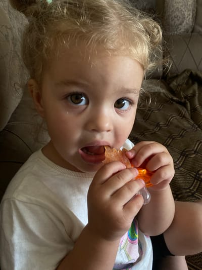 A close-up, eye-level shot shows a young child with blonde curly hair, a white t-shirt, and brown eyes. The child is sitting on a couch, holding an orange object with their hands and bringing it to their mouth. The child's mouth is open, and they appear to be eating or sucking on the object. The background is blurred, with hints of a patterned couch and blanket. The scene is indoors, likely in a home setting. The child's hair is styled in two messy buns.