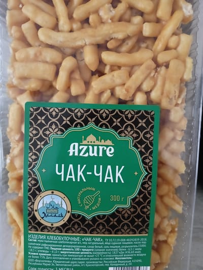 The image shows a package of Chak-Chak, a traditional Tatar and Bashkir pastry made from fried dough and honey. The package is primarily green with ornate gold and black patterns. The brand name "Azure" is prominently displayed in a white, stylized font. Below "Azure," the product name "ЧАК-ЧАК" is written in large, bold white letters. There is also a circular emblem featuring Arabic calligraphy and a crescent moon, signifying "Halal."
The Chak-Chak itself appears as a mass of golden-brown, irregularly shaped pieces, coated in a sticky glaze, likely honey. The pastry fills the transparent plastic packaging.
The text on the label provides detailed information about the product, including its composition (wheat flour, natural honey, chicken eggs, sunflower oil, sugar, salt, baking soda E500ii), nutritional information (protein, fats, carbohydrates, and calorie content per 100g), and storage conditions (temperature not exceeding +25°C, humidity not exceeding 75%). It also lists the manufacturer as "OOO «Вкуснотеевъ»" with its production address in the Mari El Republic, Russia. The expiration date is indicated as 3 months.
The scene is a close-up of the food product packaging. There are no people or specific environmental cues like time of day or weather. The location context is specified as Nizhnetroitsky, Russia, which is likely where the product is manufactured or distributed. The overall impression is one of a sweet, honey-glazed dessert.