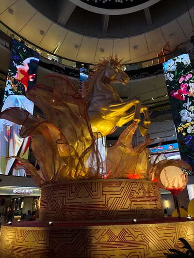 The image depicts a large, golden horse sculpture adorned with translucent, flowing fabric, situated in a multi-level indoor space. The surrounding environment suggests a shopping mall or commercial area, as evidenced by large digital screens displaying colorful imagery and advertisements. The sculpture is positioned in front of decorative panels featuring geometric patterns in gold and red. The overall aesthetic suggests an upscale, festive atmosphere. This scene is likely in Genting Highlands, Malaysia, given the context of the prompt. No people are clearly visible.