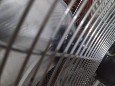 The image captures a close-up view of a metallic fan, seen through its protective grill. The foreground is dominated by the fan's silver or chrome-plated metal cage, which consists of numerous parallel, slightly curved bars running diagonally across the frame. Behind these bars, the stationary fan blades are visible, appearing to be made of a light, reflective material, possibly aluminum, showing some minor wear and reflections.

Further into the background, parts of the fan's dark-colored motor housing can be seen, secured by small bolts or screws. Through the gaps in the fan blades and motor, hints of a reddish-brown surface or object are discernible. The lighting appears soft and diffused, highlighting the metallic textures of the fan components. No specific activity is taking place, as the fan blades are clearly defined and motionless. There are no people present, and no visible text. The setting is likely an indoor environment in Gampongbaro, Indonesia, focusing on the details of this common household appliance.