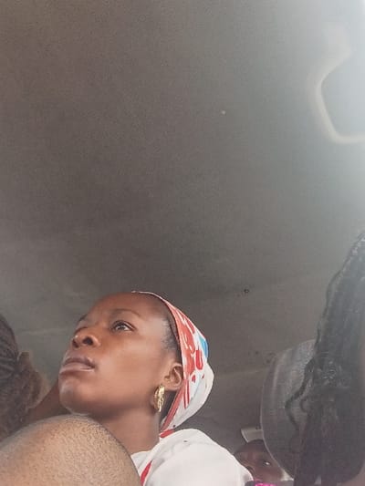 The image depicts the interior of a vehicle, likely a car or bus, containing multiple occupants. The primary subject is a young adult female positioned in the center-left, facing upwards and slightly left, with her gaze directed out of frame. She possesses dark skin and wears a white garment with a colorful headscarf, predominantly white with red and blue patterns, covering her hair. A gold-colored, ornate earring is visible in her right ear. Her left shoulder and upper arm are prominent in the foreground. To the left and slightly behind her, another individual's dark, possibly braided hair is visible. To the right, partially obscured, is another person with long, dark braided hair, and a child's partially visible face, featuring one eye and forehead, is discernible below and to the right of the main subject. The vehicle's ceiling is a light grey/beige fabric. Natural light appears to illuminate the scene from the right side. The exact location within Itobe, Nigeria, is not discernible from the visual information provided.