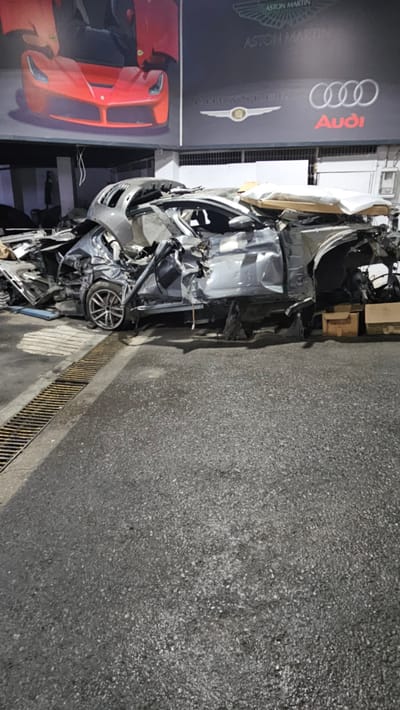 A severely damaged grey vehicle is parked on an asphalt surface, which appears to be a street or parking lot in Ataşehir, Turkey. The car is mostly obliterated, with its body panels crumpled and torn. Various parts of the vehicle are scattered around it, including what looks like a large piece of metal resting on top of the wreckage. In the background, a mural of a red sports car is visible on a wall, alongside logos for Aston Martin, Chrysler, and Audi. The scene suggests a significant car accident has occurred.