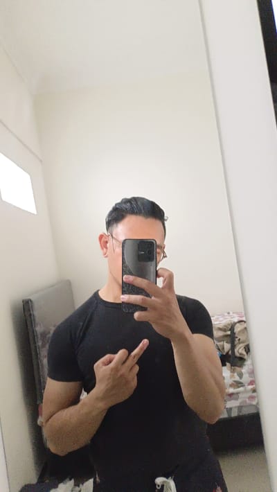 A man is taking a selfie in a mirror. He is wearing a black t-shirt and has his middle finger extended. He is holding a black smartphone in his right hand and is looking at the camera. The room has white walls and there is a bed with a headboard in the background. This image was taken in Magelang, Indonesia at the coordinates: latitude -7.479, longitude 110.222. The image is taken from the waist up. The lighting in the room is bright, and the man's face is illuminated. The background of the image is out of focus, but a bed and a floral patterned blanket can be seen. The man's left hand is obscured from view. The room is cluttered with personal belongings.