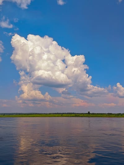 The image depicts a wide, calm river flowing through a lush green landscape under a bright blue sky dotted with fluffy white clouds. The river's surface reflects the sky and the distant treeline. The horizon is marked by a dense band of green foliage, suggesting a forested area. In the distance, a thin line of trees borders the river. The overall scene is tranquil and natural, evoking a sense of peace and vastness. The location is identified as the city of Yeniseysk, Russia.