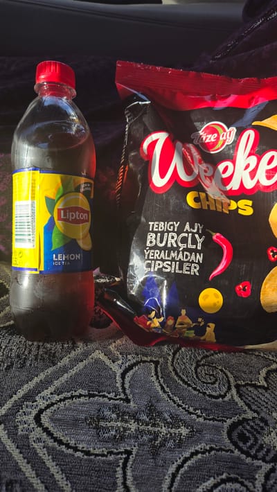 An outdoor, low-angle shot shows a bottle of Lipton Ice Tea and a bag of "Weke" chips on a patterned textile. The bottle, with a red cap, is on the left and appears to be sweating. Its label is yellow and blue, featuring an image of lemons and the Lipton logo. The bag of chips, mostly black with red and white lettering, is on the right. The text on the bag is in a non-English script. The background is dark and blurry, suggesting a car interior or shaded outdoor setting. The scene is set in Sakarçäge District, Turkmenistan.