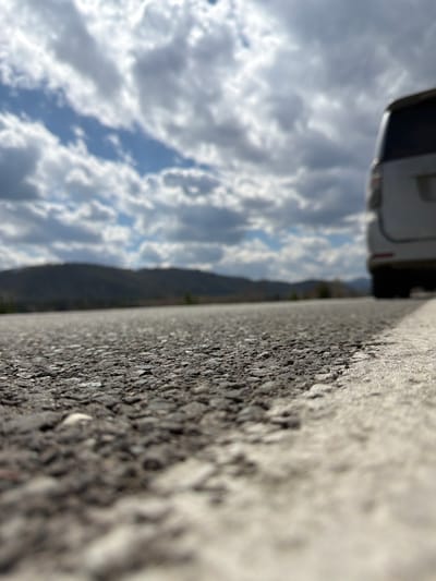 The image, taken at a low angle near a road surface, features a close-up view of asphalt pavement with a white painted road marking. The asphalt texture is coarse. In the background, there is a mountain range and a sky filled with clouds. A vehicle, possibly a camper or van, is partially visible in the lower right corner. Based on the city specified, the location is likely somewhere near Сизая, Russia, but there are no specifics. There are no people shown in the scene.
