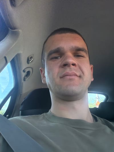 A man is in the driver's seat of a car. He has short dark hair, and is wearing a light gray T-shirt and a seatbelt. He appears to be taking a selfie. Through the car window, there are buildings and trees. Given the prompt about the location, it can be inferred that this image was taken somewhere within the city of Tulovo, Bulgaria. There are no specific details to identify a particular location within the city.
