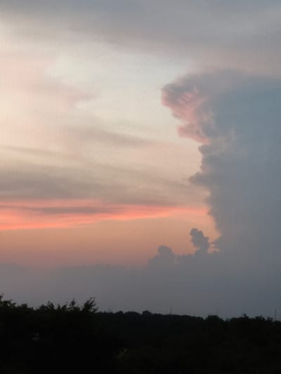This image depicts a sunset sky over a dark, silhouetted landscape of trees and distant structures, likely in Kerpen, Germany. The sky is dominated by dramatic clouds with varying colors. On the right, a large, dark gray cloud formation rises ominously, tinged with pink and purple on its left edge, suggesting the last rays of sunlight are catching its texture. To the left of this imposing cloud, the sky transitions into softer hues of pale pink and orange, forming distinct horizontal bands of color, indicative of a sunset. Further to the left and higher up, the sky fades into a hazy, light gray. The lower portion of the image is filled with a dense, dark silhouette of trees, creating a stark contrast with the illuminated sky above. There are faint outlines of what appear to be structures or distant lights visible through the trees on the horizon, hinting at an urban or semi-urban setting. The overall mood is dramatic and serene, capturing the fleeting beauty of a sunset. The time of day is clearly evening, and the weather appears to be clear with the exception of cloud formations. No text is visible in the image.