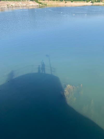 This is a photograph of a body of water, likely a lake or reservoir, viewed from above. The water is divided into two distinct sections, with a lighter, calmer blue on the left and a darker, murkier blue on the right, separated by an area with visible aquatic vegetation. The left side of the image shows a shoreline with a paved walkway or road and green vegetation. Several small white objects, possibly buoys or distant boats, are scattered on the lighter blue water.
A prominent feature on the right side of the water is a large shadow cast by an unseen object or structure, creating a stark contrast with the illuminated water. Within this shadow, the silhouettes of two people are visible, appearing to be standing and possibly waving or gesturing. A thin pole, likely part of the structure casting the shadow, extends upwards through the silhouettes.
The overall scene suggests a sunny day, given the clear shadows. The time of day appears to be midday or afternoon, judging by the angle and length of the shadow. The presence of the shoreline, vegetation, and water suggests an outdoor recreational or natural setting. The people in the shadow are the only clearly discernible human subjects. There is no visible text in the image. The location context provided is Korzhevo, Moldova.