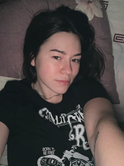 A young woman with dark hair and light skin is lying on a pillow with a floral print. She is wearing a black t-shirt with a white graphic design and the words "BORN TO BE FREE." A silver chain necklace is visible around her neck. Her arm is raised, revealing a faint tattoo. She is gazing at the camera with a slight smirk. The lighting is dim, casting shadows across her face and the surrounding area.