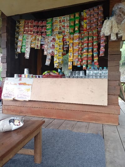 The image shows a small, open-air shop in the town of Pangalengan, located in the Bandung Regency of West Java, Indonesia. The shop is constructed of wood and has a wooden roof. It is adorned with numerous colorful packets of snacks and drinks. There are also some canned goods and bottled drinks on the counter, as well as some empty glasses and cups. On the wooden counter in front of the shop is a sign printed with an Indonesian company logo and message. A wooden bench with a single, wrapped snack on its surface is visible in the foreground. The shop appears to be closed, as there are no customers or vendors present, and a gray mat is positioned on the wooden floor to the right of the bench. The shop is located within a larger, more complex structure, as one can see a wooden post and an opening to another room. The shop is situated in a rural location, surrounded by lush greenery. The surrounding area is well-maintained and clean. The coordinates are approximately -7.233° latitude, 107.537° longitude.