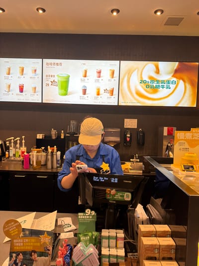 The photo shows the interior of a Starbucks store, likely located in Yuhuatai District, China, based on the presence of Chinese characters and references to promotions specific to that region. A Starbucks employee wearing a blue uniform and a khaki cap is holding a smartphone with a brightly colored case near the point-of-sale terminal.
To the left of the employee is a digital menu board displaying a variety of drinks with prices listed in Chinese currency. The menu includes classic coffees, barista favorites like matcha lattes, and popular picks such as coconut velvet and oat lattes. Advertisements for promotional offers are also present on the board, one of which highlights a product with 20 grams of native milk protein.
In front of the employee are the payment terminal, and multiple stacks of Starbucks-branded wafer roll boxes. Various packaged food items, including packets of "original flavor nuts" and "sugar-free mint flavor gum" are visible to the right of the terminal. Promotional material advertising a "Buy 3 get 1 free" offer is also displayed.