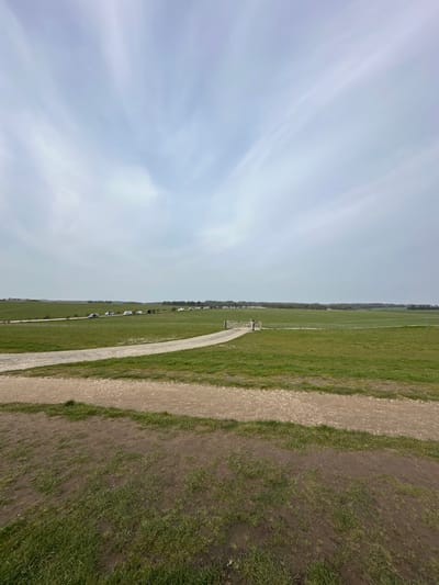 The image depicts a wide-angle view of a rural landscape near Amesbury, United Kingdom, under an overcast sky with wispy cloud formations. The foreground consists of mixed grass and dirt patches. A light-colored dirt or gravel path curves from the lower center-right into the mid-ground, where it approaches a wooden five-bar gate.
Beyond the gate, expansive green fields stretch towards a distant tree line. Along a slightly elevated, curving track or road in the mid-distance, a linear arrangement of approximately 15-20 vehicles (appearing to be cars, vans, and potentially caravans/RVs) is visible. Further beyond these vehicles, nearer to the distant tree line, a cluster of white tents or temporary structures is discernible.
One human figure, appearing to be an adult, is visible standing near the wooden gate on the dirt path. The overall environment is open grassland, suggesting agricultural or common land, with diffuse lighting typical of an overcast day.