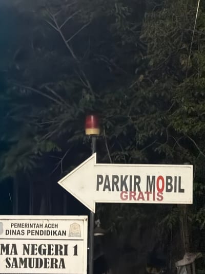 The image displays a nighttime or dusk scene featuring two informational signs and a warning light against a dense, dark green foliage background. A prominent white arrow-shaped sign points left, bearing the Indonesian text "PARKIR MOBIL" (Car Parking), with the word "GRATIS" (Free) overlaid in red, partially obscuring the letter 'O' in "MOBIL." Directly above this sign, a red and amber/yellow rotating beacon or warning light is mounted on a pole. Below the arrow sign, a rectangular white sign is visible, displaying "PEMERINTAH ACEH DINAS PENDIDIKAN" (Government of Aceh, Education Department) and "MA NEGERI 1 SAMUDERA" (Public Senior High School 1 Samudera), accompanied by an institutional emblem. The vegetation in the background consists of trees or bushes, indicating an outdoor, possibly semi-urban or rural setting. No individuals or active interactions are present. The signs confirm the location is at or near Public Senior High School 1 Samudera, situated in the Samudera district of North Aceh Regency, Aceh Province, Indonesia, which includes the specified area of Beurandang.