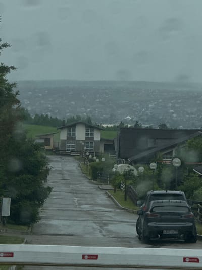 The image shows a view of a residential area in Kuda, Russia, on a cloudy day with visible raindrops on the camera lens. In the foreground, a white barrier with red "MOTORS" logos is visible. Beyond the barrier, a wet asphalt road curves uphill, flanked by greenery and trees on the left and several houses on the right. The houses appear to be modern, multi-story structures with dark roofs and light-colored siding. One prominent house in the middle ground has a gray and white facade with multiple square windows. Further up the road, another house with a dark roof is visible. In the mid-ground, two cars are parked on the right side of the road: a dark SUV and a dark sedan with a large rear spoiler. The sedan has a license plate with Cyrillic characters and numbers "H367EO 128". In the background, a vast expanse of a city or town stretches across the horizon, partially obscured by atmospheric haze and low-lying clouds. The overall impression is one of a somewhat overcast, suburban or semi-rural setting.