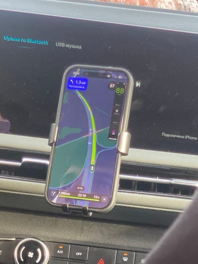 An iPhone is mounted to a car's air vent, displaying a map app with navigation. The route guides towards Rostovskoe sh, approximately 1.3 km away. The car is connected to the iPhone. Other information visible includes a speed of 87-88, distance to destination (1290km), arrival time (23:10), time until arrival (12h). Media can be played via Bluetooth or USB.  A/C and OFF button on the car's dashboard are also visible.