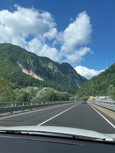 The image depicts a road trip through a mountainous region with a clear blue sky and scattered white clouds. The road is paved with white lane markings and flanked by metal guardrails. On either side of the road, lush green trees and steep, rocky mountains rise dramatically. One mountain face shows a visible landslide scar. In the distance, a tunnel entrance is visible on the right side of the road, and a few cars are present on the highway. The viewpoint is from inside a vehicle, looking forward. The location is not Monastyr, Russia, as this image depicts a mountainous landscape inconsistent with the known geography of Monastyr.