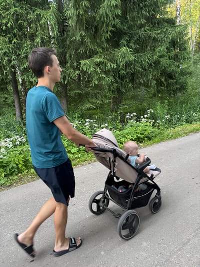 A young man is pushing a stroller with a baby in it down a paved path. The man is wearing a teal t-shirt and black shorts. He is wearing black slides. The baby is wearing a light blue outfit and white socks and is lying down in the stroller. The stroller is beige and black. The path is lined with lush green trees and plants. There are some small white flowers growing in the greenery. The sun is shining, casting light and shadows on the scene. The time of day appears to be daytime. The location is likely a park or a suburban area with walking paths, possibly in Шевлягино, Russia. The overall mood seems peaceful and pleasant, with the man enjoying a walk with the baby. There is some visible text on the man's slides, which appears to say "SANS".