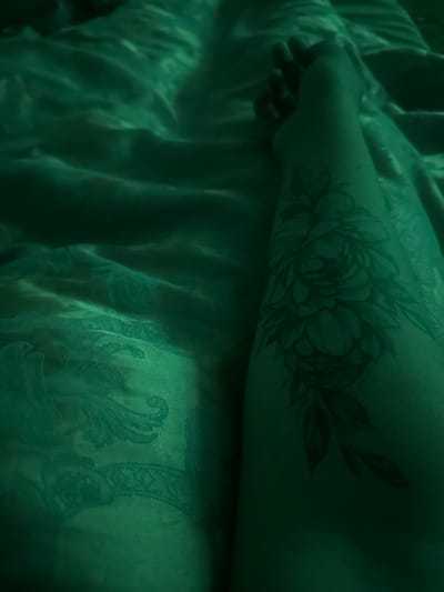 The image shows a close-up of a person's arm resting on a green, textured fabric, possibly a bedspread or blanket. The fabric has an embossed or patterned design visible in the light. A tattoo of a floral design, featuring a large bloom and leaves, is visible on the person's forearm. The lighting is dim and casts a green hue over the scene, obscuring some details. There are no visible indications of the location being in Gavrilov Posad, Russia, or any urban environment.