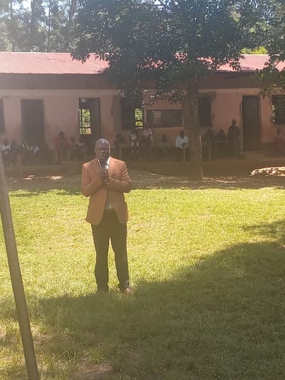 A full-length outdoor image captures a scene in Idakho South ward, Kenya, featuring a man addressing a group in front of a building. In the foreground, a dark-skinned man, appearing middle-aged to elderly with short grey hair, stands centrally, facing slightly right. He is dressed in a light brown suit jacket, white shirt, dark trousers, and reddish-brown shoes. His hands are clasped at chest level, holding a dark object. Approximately 12 to 15 individuals are visible in the mid-ground, seated and standing in front of a single-story building. This building has a light pink facade, a reddish-brown corrugated metal roof, and multiple open doorways and window apertures. A large tree with dense dark green foliage dominates the upper center and right, casting shadows across the foreground's green grass. Additional trees are visible in the distant background. A dark vertical pole is partially visible in the lower left foreground. The scene is illuminated by natural light, indicating a sunny outdoor setting.