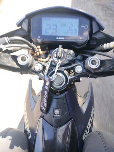 An action shot from a first-person perspective shows a motorcycle rider's point of view, with the handlebars and digital dashboard of a Suzuki motorcycle prominently featured. The rider's legs, clad in dark pants, are visible at the bottom of the frame. The dashboard displays a speed of 23 km/h, the time is 8:30, and the RPM gauge is active. A keychain with a black lanyard featuring white text, possibly "VAULT," hangs from the ignition. The motorcycle itself is dark blue, with "SUZUKI" branding visible on the fairing. The background is blurred, suggesting movement and an outdoor environment, likely a road. While the image provides a close-up of the motorcycle's controls, it does not contain enough information to definitively place it within Kidapawan City, Philippines, or any specific urban environment.