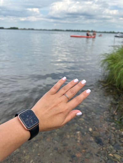 A person's hand, with white nail polish and a ring, is in the foreground, wearing a rose gold smartwatch with a black band. In the background, on a lake, two people are on a red floatation device. The scene is set in Tasheba, Russia, with water, pebbles, and grass visible.