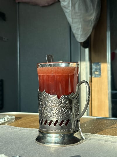 A clear glass containing a red, opaque beverage with a frothy top layer is positioned in a silver-colored, ornate metal cup holder featuring a handle and decorative embossing. A small metal spoon rests inside the glass. The glass and holder are situated on a light-colored wooden table surface partially covered by a white, light gray cloth. In the background, dark gray vertical paneling is visible on the left, while a lighter brown vertical panel or door frame with metallic fixtures is present on the right, illuminated by sunlight casting a harsh shadow. A light-colored, possibly plastic, bag or object hangs suspended in the upper right background. The setting is consistent with the interior of a train car, specifically observed during transit in Volkhov, Russia. No people are visible.