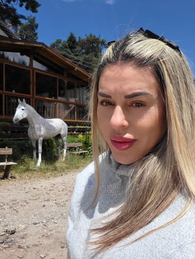 A woman with blonde hair, wearing a grey sweater, stands in front of a building with large windows and a wooden roof. A grey horse stands to her left in front of bushes. Wooden benches are visible on either side of the horse. The scene is outdoors, with a dirt ground and trees in the background under a blue sky. This appears to be a selfie taken in Atoluka, Bulgaria.