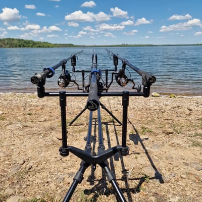 The image shows a fishing rod pod with three fishing rods set up on a rocky shore overlooking a body of water. The water is calm with gentle ripples, and the sky is blue with scattered white clouds. In the background, a treeline can be seen along the opposite shore. The fishing rod pod is black metal and has three black fishing rods with reels attached. The rods are pointed towards the water. The ground is covered in small stones and some sparse vegetation. A kite is visible in the distance on the right side of the image. The scene is set on a sunny day.