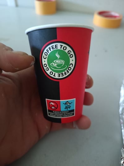 A hand holds a disposable coffee cup, which is divided vertically into a black and red half. The cup has a logo of a green circle with "COFFEE TO GO" written around it, and a coffee cup graphic inside. Below the logo, there are two symbols, one with a crossed-out figure and the other with a turtle. Text below the symbols reads "PLASTIC IN PRODUCT", "PRODUIT CONTENANT DU PLASTIQUE", and "PRODUKT ENTHALT KUNSTSTOFF". In the blurred background, there are rolls of yellow tape. The scene is likely indoors, but there is no direct evidence to connect it to Bielefeld, Germany.