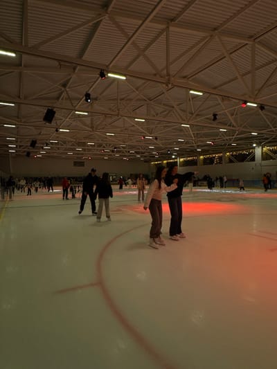 The scene is an indoor ice skating rink located in the city of Terk, Russia. The facility features a high, vaulted ceiling composed of white corrugated panels and a network of exposed metal trusses. Illumination is provided by numerous fluorescent strip lights and spotlights, some of which project a red light onto the ice surface. The ice itself displays a curved red demarcation line. Multiple individuals are observed engaging in ice skating. In the foreground, three individuals are positioned center-right. One, wearing a light-colored long-sleeve top and dark trousers, skates towards the right. Adjacent to them, another individual in a dark long-sleeve top and dark trousers extends their right arm in a forward pointing gesture while also skating rightward. A third individual, in dark attire, is partially visible to the right. All three are equipped with ice skates. Further into the rink, numerous other skaters are present, including a male in a dark hoodie and dark pants, and an individual in a dark jacket and light pants. Some distant figures appear to be utilizing skating aids. The background wall features decorative string lights.