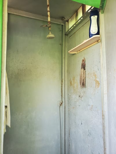 An outdoor shower stall is depicted, constructed from what appears to be corrugated metal panels painted a faded grey. The shower head, made of brass and showing signs of rust, hangs from a corroded metal pipe extending from above. A towel, a light beige color, is draped over a pipe on the left side of the stall. On the right wall, a small, light-colored wooden shelf holds a blue bottle of shampoo or body wash. Below the shelf, a woven object, possibly a loofah or washcloth, hangs from a nail. The walls of the stall exhibit rust stains and peeling paint, suggesting age and exposure to the elements. The surrounding environment suggests an outdoor or semi-enclosed space, with glimpses of green structures and bright light, possibly indicating daytime. The scene is devoid of people, and no actions or interactions are visible. The overall impression is one of a functional, utilitarian, and weathered outdoor shower.