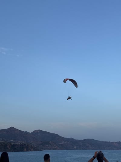 A paraglider with a red and white wing is flying in a clear blue sky over the sea and mountainous coastline. The paraglider has a small frame with a person sitting in it. Below the paraglider, the blue water of the sea stretches to the horizon where dark, tree-covered mountains rise. The mountains appear to be part of the Nerja coastline in Spain. In the foreground, three people are visible, two facing away from the camera and one facing towards it. One person on the right is holding a smartphone, possibly taking a picture of the paraglider. The lighting suggests it is late afternoon or early evening.