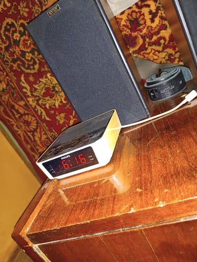 A close-up, angled shot shows a digital alarm clock with red numerals displaying "6:16" on a polished wooden surface. The clock, a "PHILIPS" brand, is white with a black display. To the left of the clock is a black speaker with a gold "SVEN" logo. The background features a red and gold patterned carpet and a yellow wall. A white cable extends from the clock. A dark-colored rolled-up object, possibly a belt, is visible behind the speaker. The scene appears to be indoors.