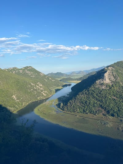 The image depicts a wide, winding river flowing through a valley flanked by lush, green, tree-covered mountains. The sky is a clear blue with scattered white clouds. The river reflects the sky and the surrounding landscape. The banks of the river are lined with dense vegetation, including reeds and smaller trees. In the distance, more rolling hills and mountains are visible, creating a sense of depth. The overall scene is one of natural beauty and tranquility. The location is specified as Rvaši, Montenegro.